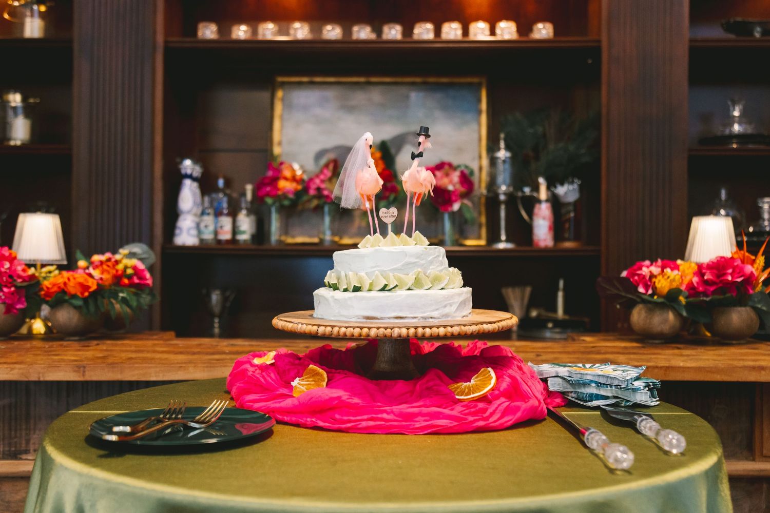 White wedding cake with flamingo toppers displayed on gold stand with pink and green table decor.