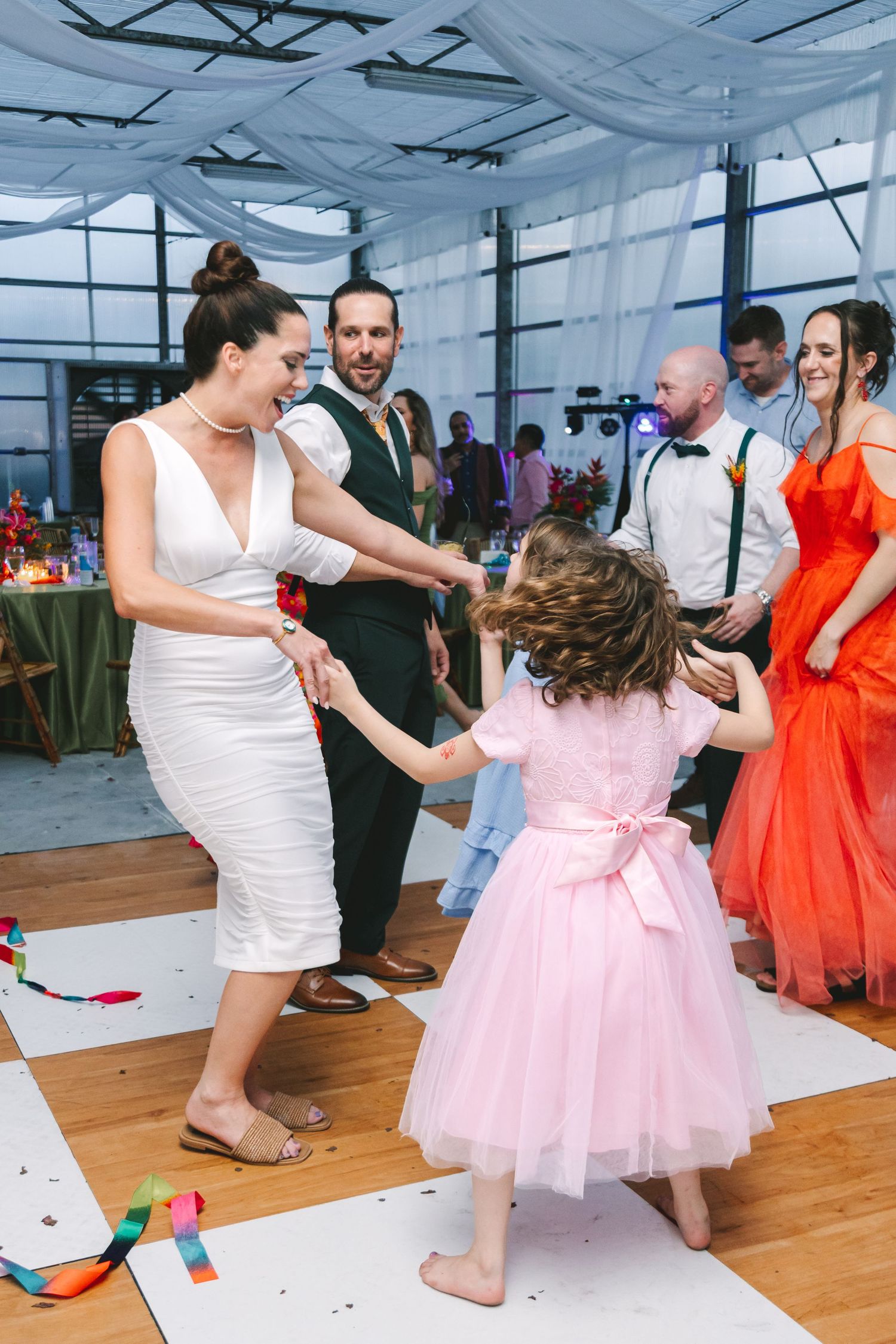 Bride in white dress dances joyfully with young flower girl in pink tutu at wedding reception.