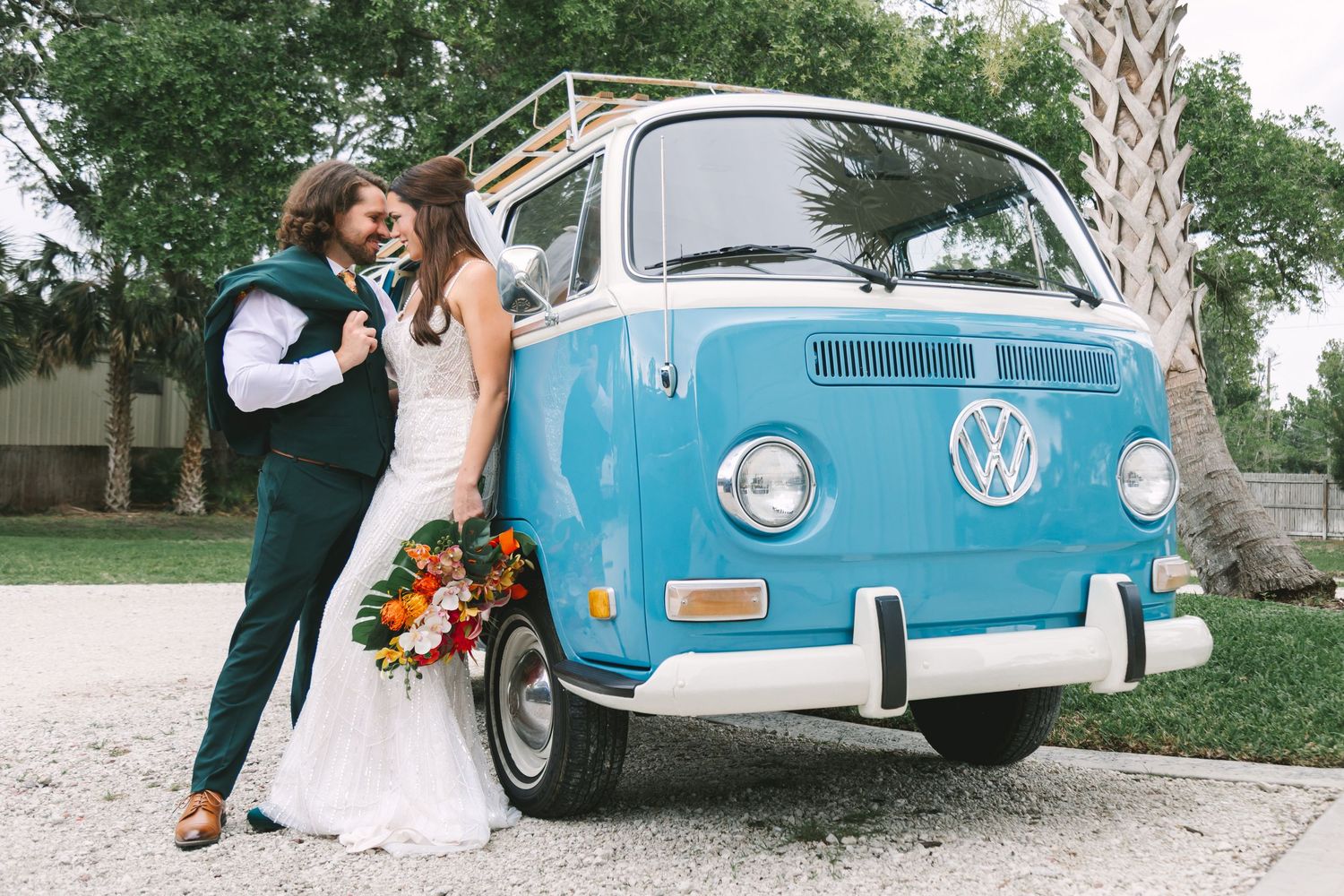 Bride and groom posing in front of a vintage blue VW bus, bride holding a colorful bouquet on their wedding day.