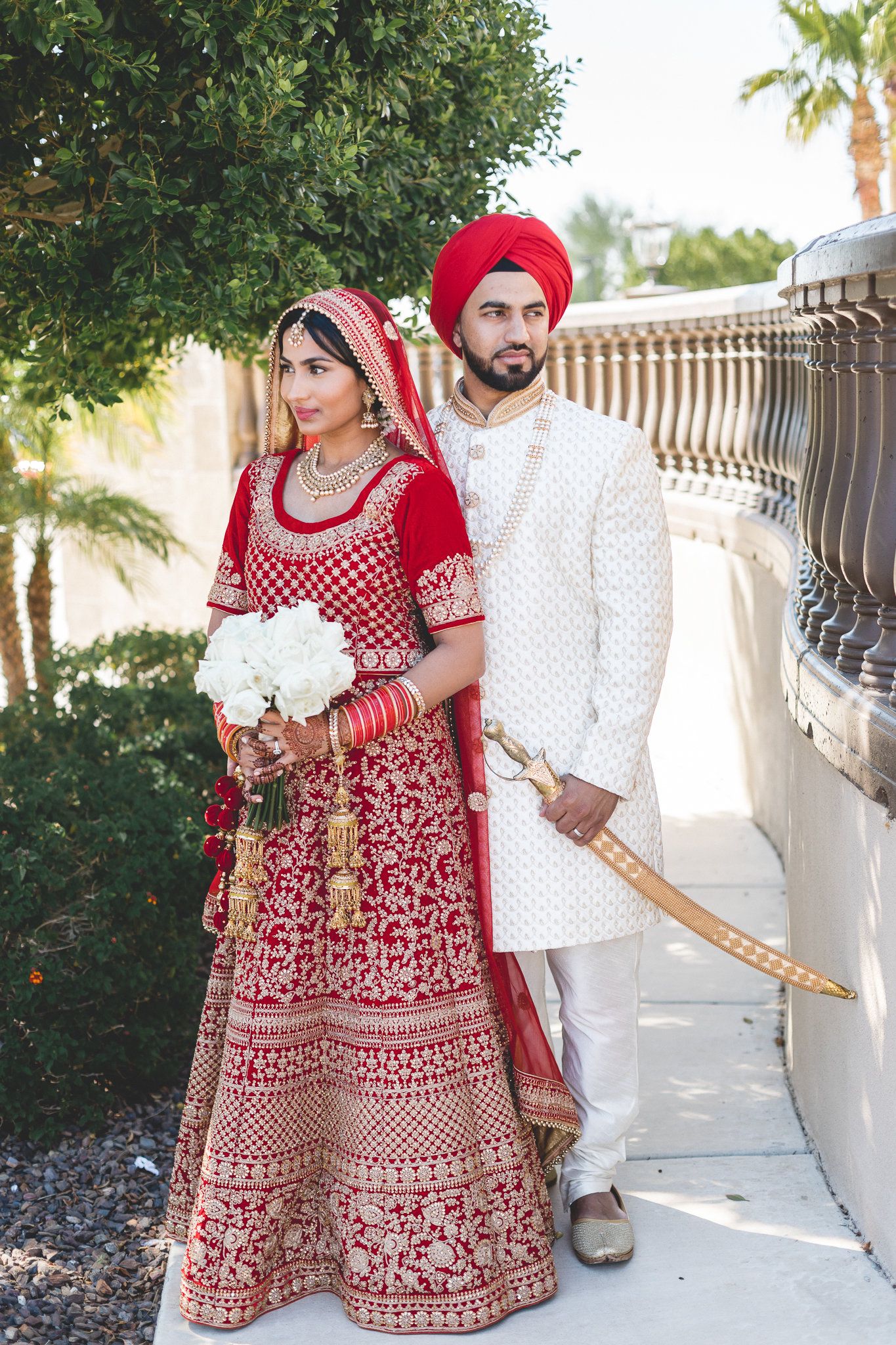 Sikh bride in red lehenga holding white bouquet with groom in white sherwani holding ceremonial sword.