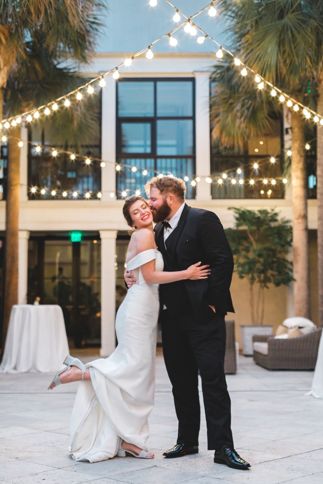 Joyful bride and groom dancing in a courtyard under glowing string lights at night.