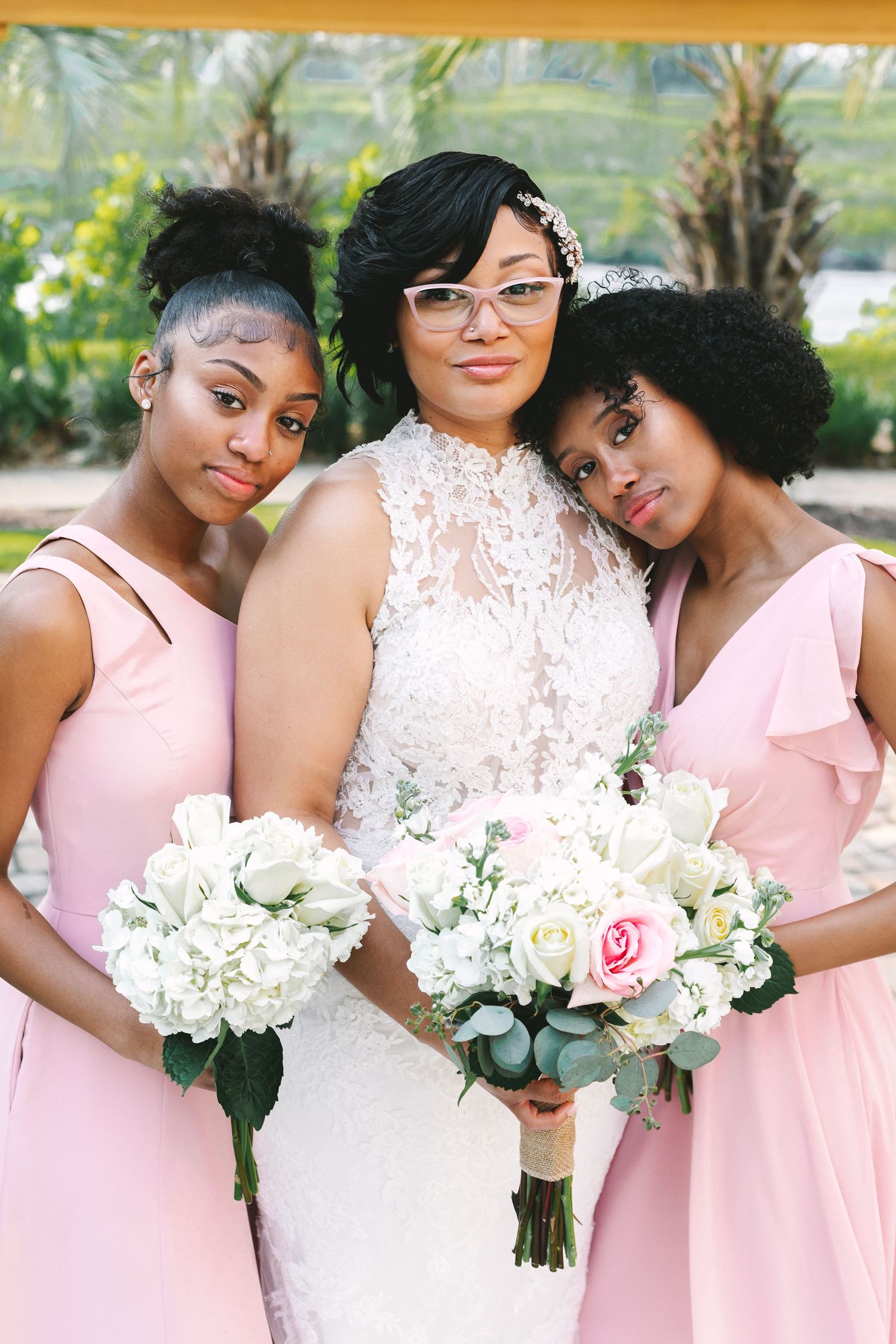 Bride wearing lace gown and glasses poses with two bridesmaids in pink dresses holding white floral bouquets.