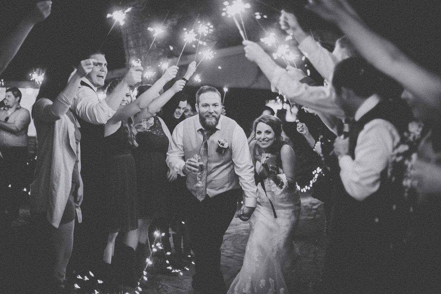 Bride and groom celebrate with sparklers surrounded by cheering wedding guests in black and white photo.