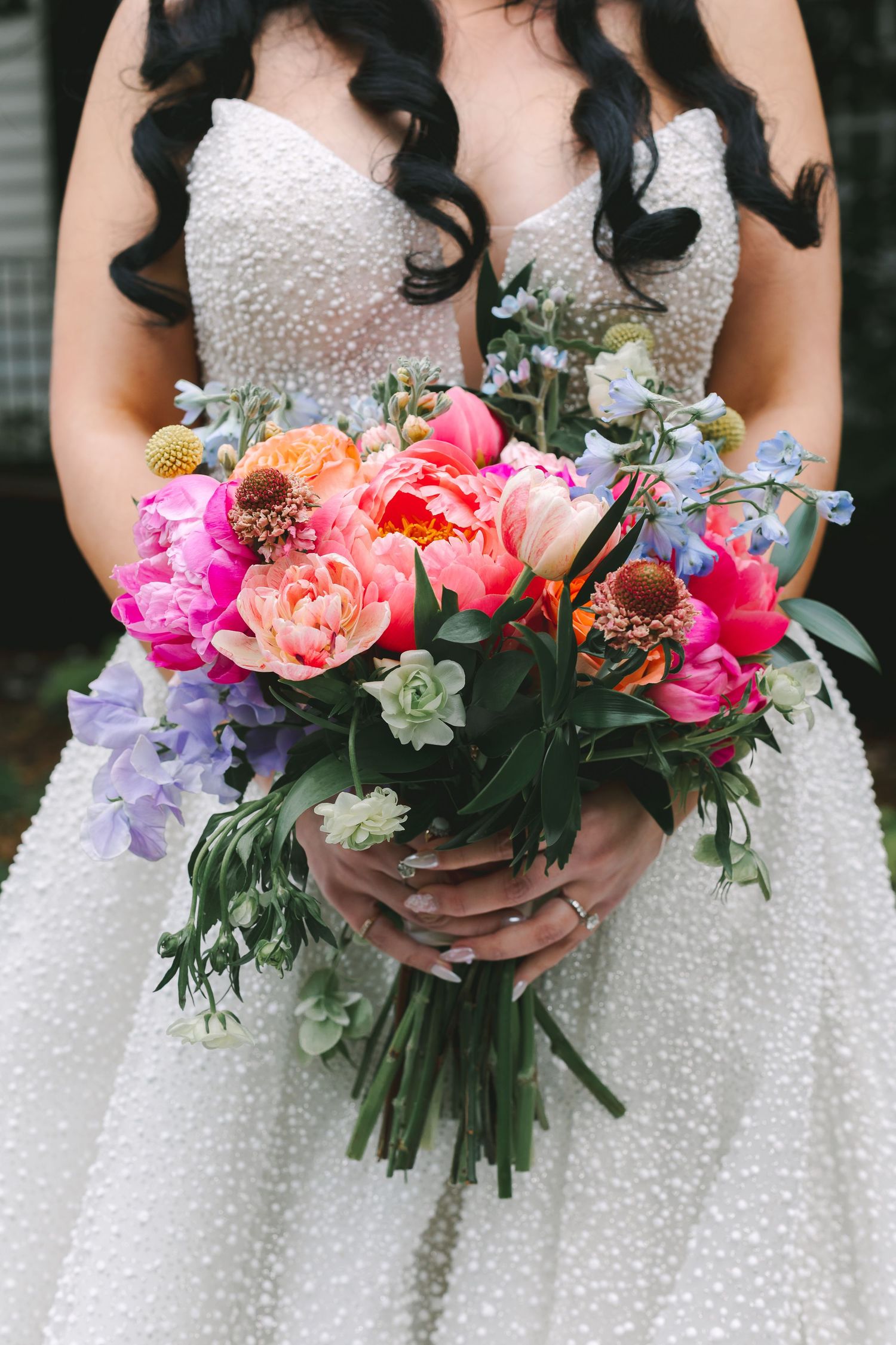 Bride in pearl wedding gown holding vibrant bouquet of pink peonies, tulips, and blue wildflowers.