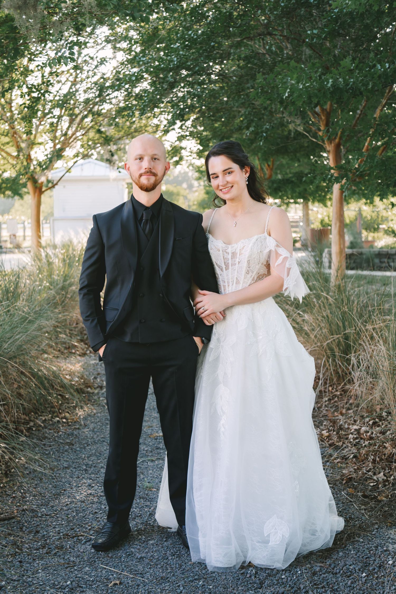 Smiling bride and groom pose together outdoors, she in a white tulle gown, he in a black tuxedo.