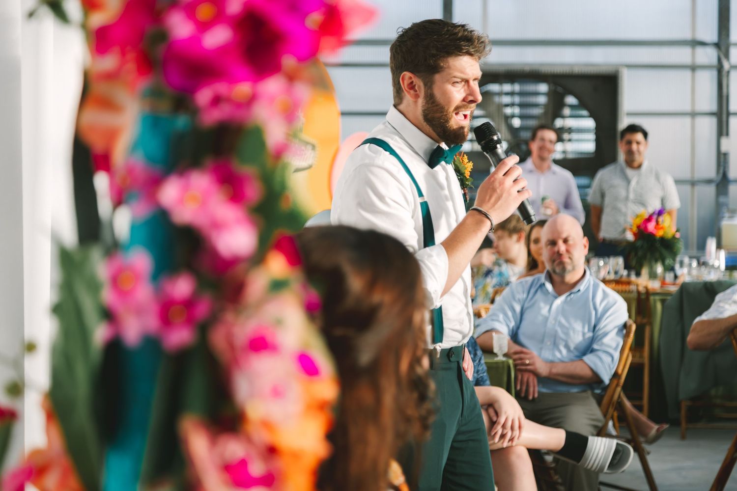 Groomsman in suspenders and bow tie giving a speech at a colorful wedding reception.