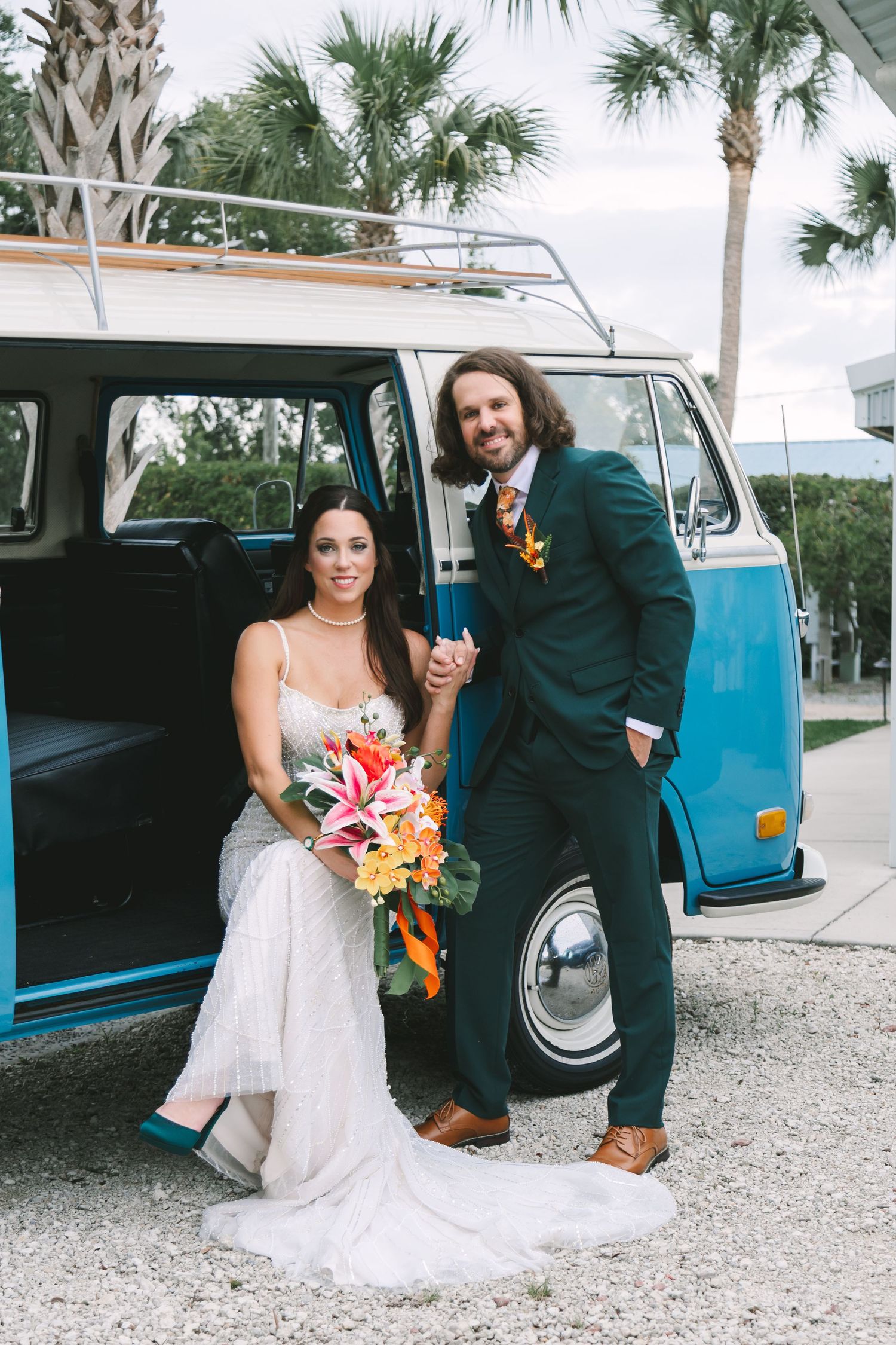 Bride and groom posing by a vintage blue VW bus, bride holding tropical bouquet, groom in teal suit, outdoor wedding photo.