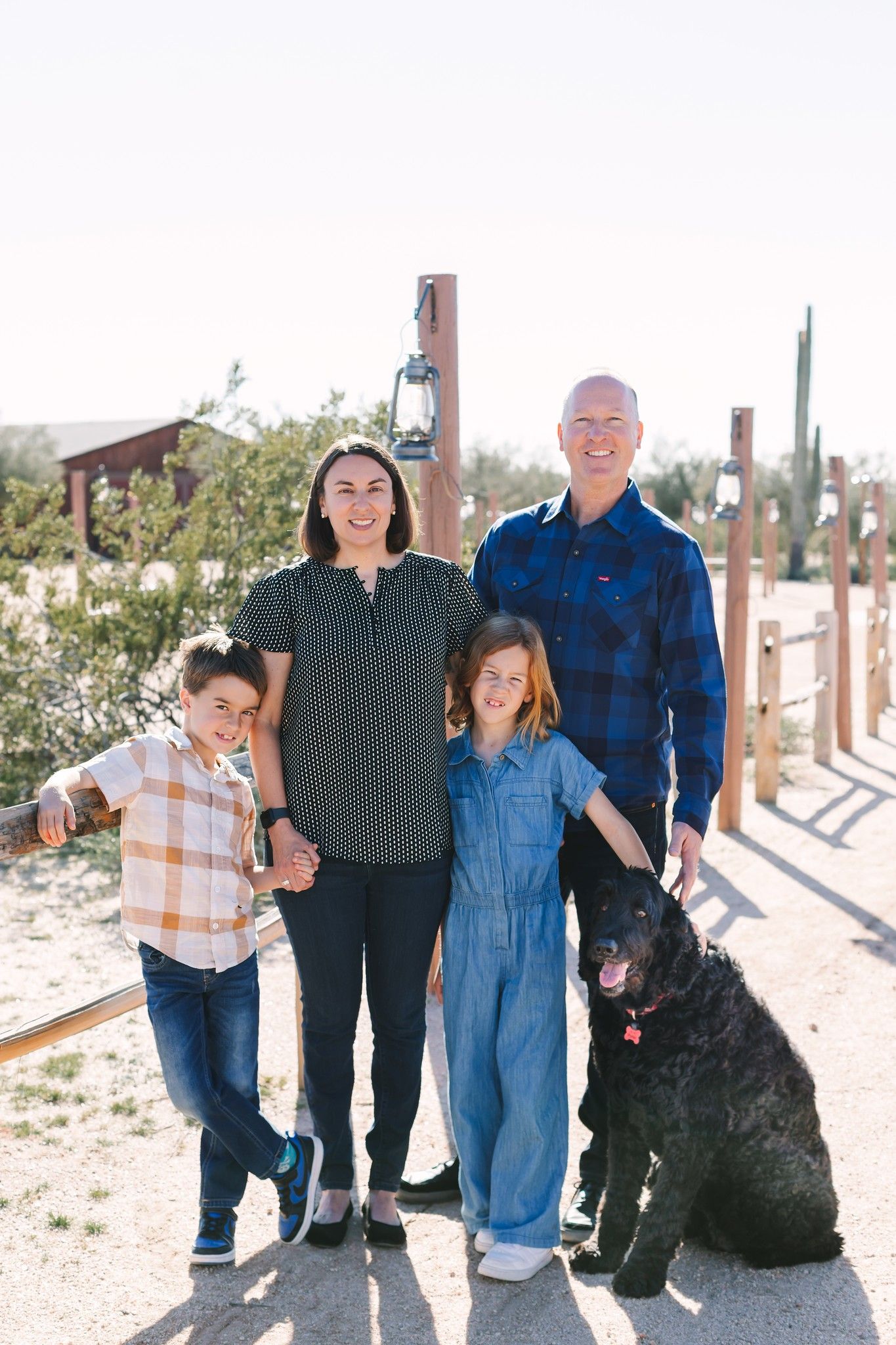 Family of four with black dog posing on desert path with saguaro cacti in Arizona sunshine.