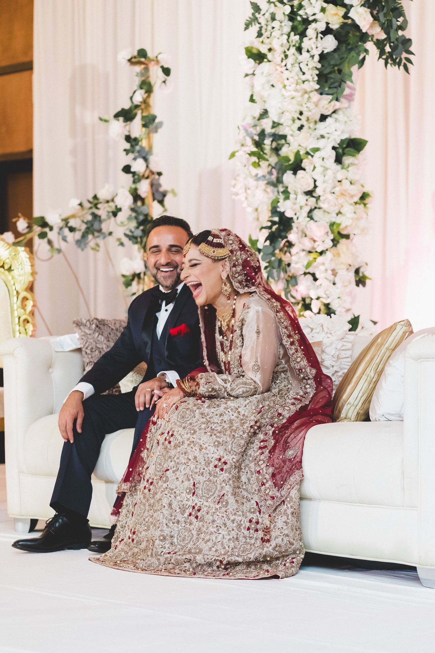 Laughing South Asian bride and groom in traditional wedding attire seated on white sofa with floral backdrop.
