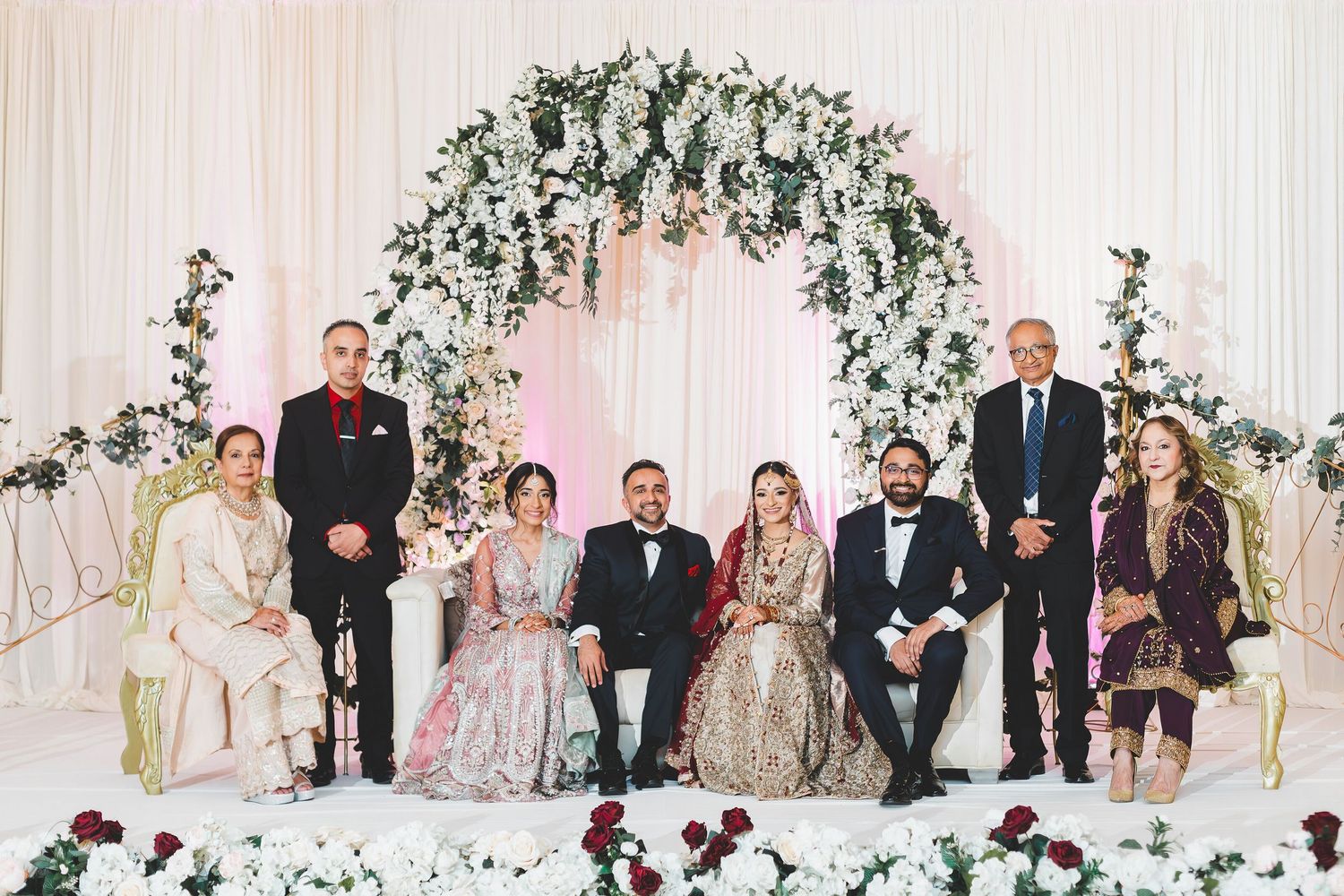 South Asian wedding couple with family posing under floral arch with white roses at reception ceremony.