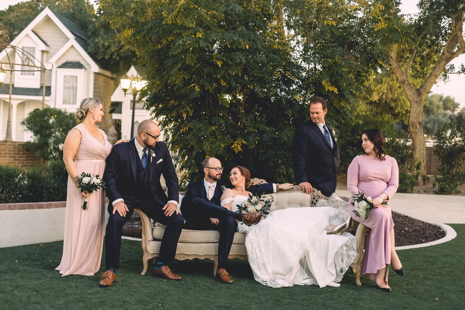 Wedding party posing casually on vintage sofa outdoors with bridesmaids and groomsmen in garden.