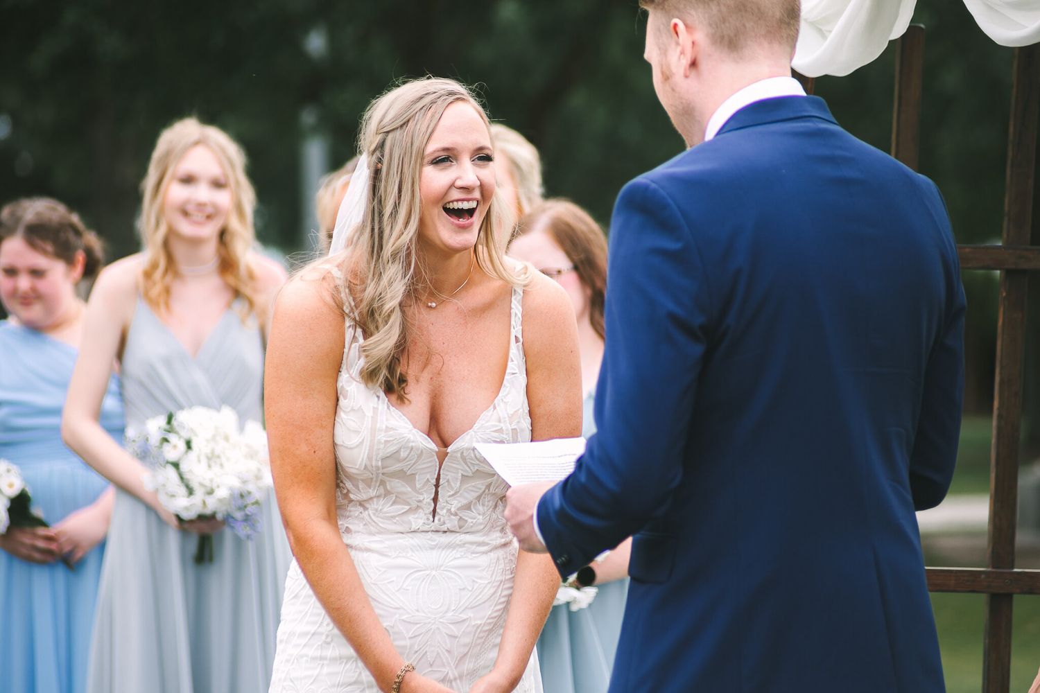 Joyful bride laughing during outdoor wedding ceremony with bridesmaids in blue dresses.