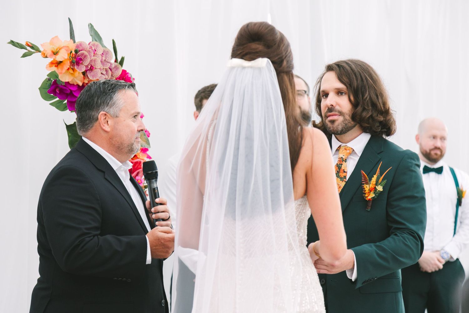 Officiant with floral crown speaks at outdoor wedding ceremony as bride in veil and groom in dark suit exchange vows.