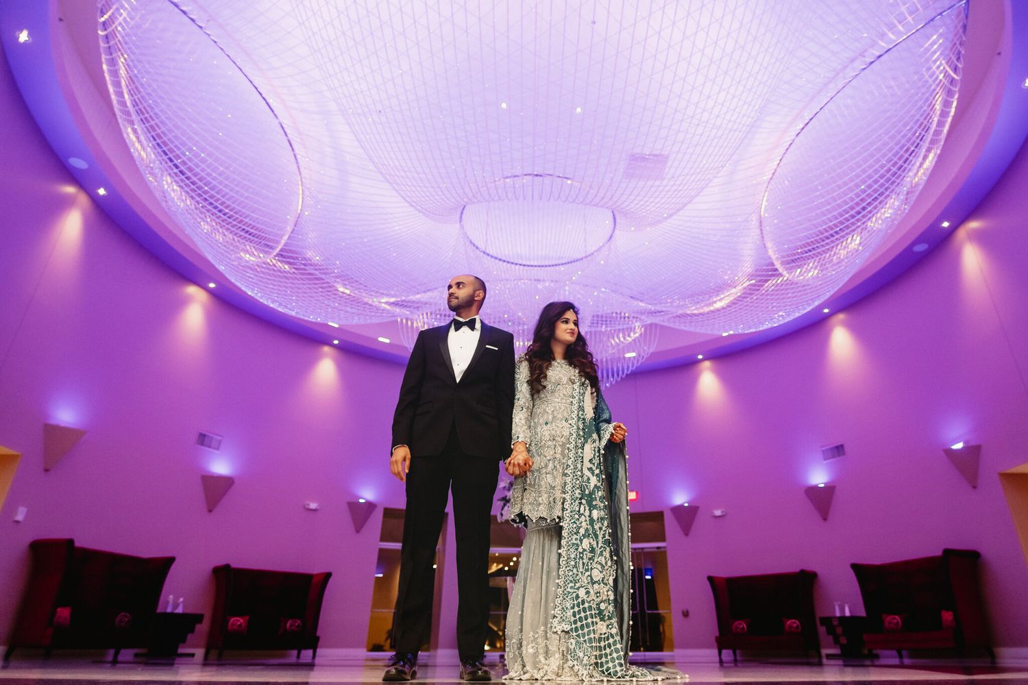South Asian wedding couple standing elegantly under a purple-lit ornate ceiling in a grand banquet hall.