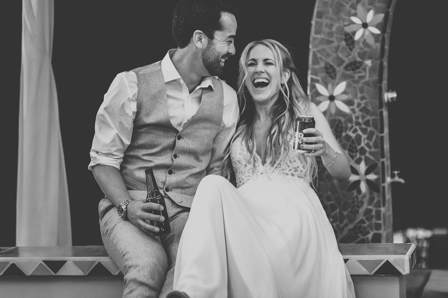 Black and white photo of laughing bride and groom sharing a drink at their wedding reception.