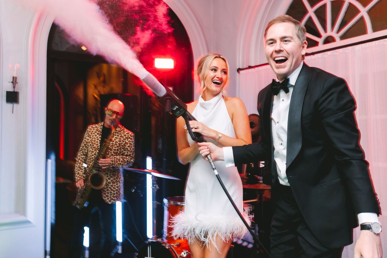 Joyful bride in white feathered dress sings into microphone with groom and saxophonist at lively wedding reception.