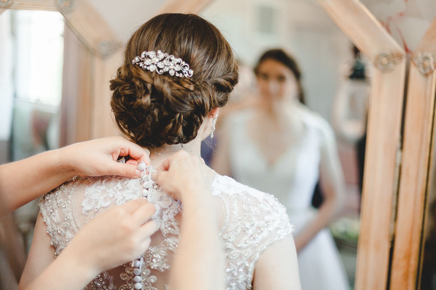 Bridesmaid buttoning the back of a bride's lace wedding dress while she looks in a mirror.