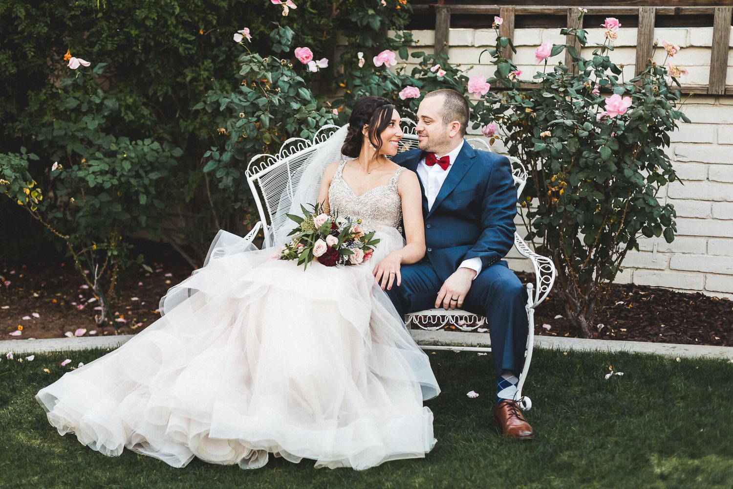Bride in layered tulle gown and groom in blue suit sitting on garden bench.