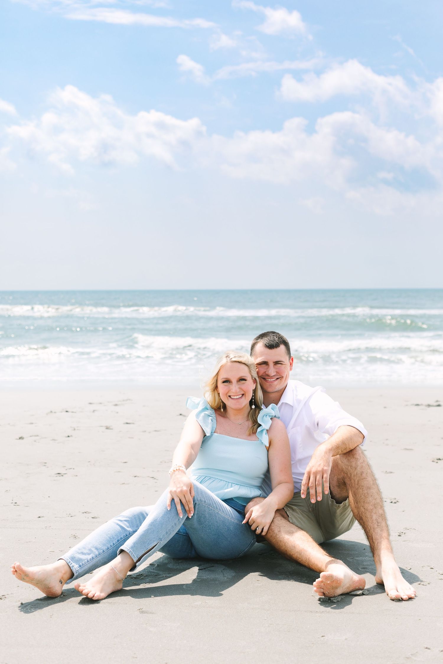 Happy couple sitting barefoot on sandy beach with ocean waves and blue sky behind them.