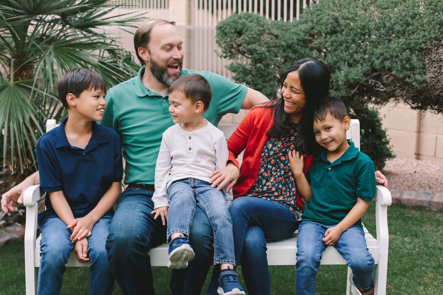 Joyful family of five laughing together on a white bench in a sunny outdoor garden setting.
