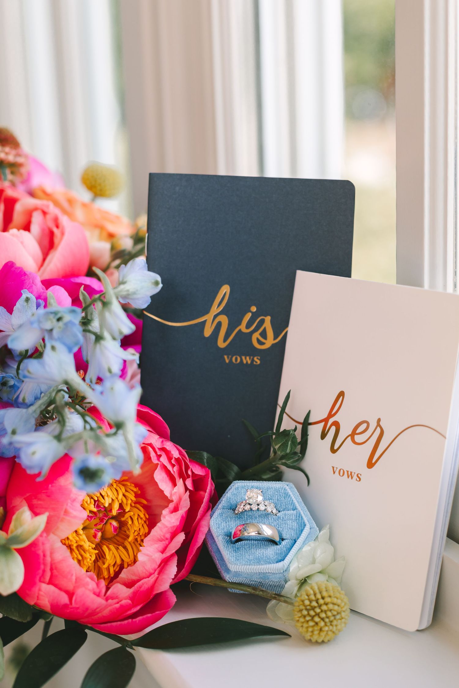 Wedding flat lay with his and her vow books, rings in a blue velvet box, and colorful flowers.