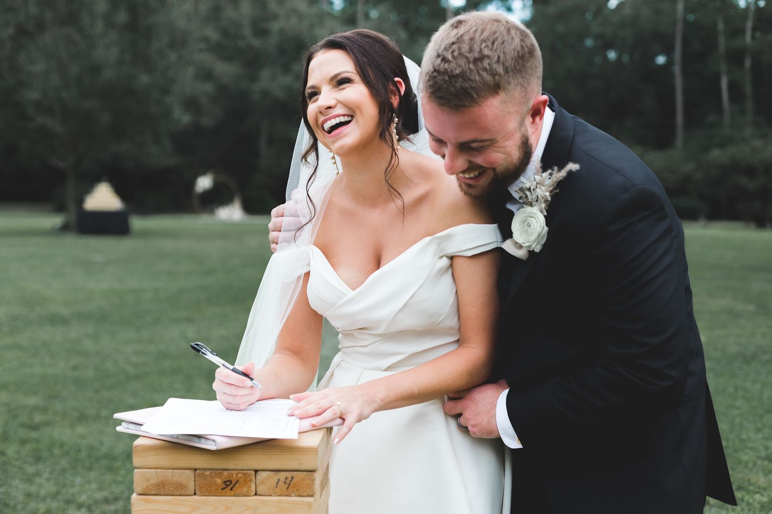 Laughing bride signs marriage certificate as groom embraces her outdoors.