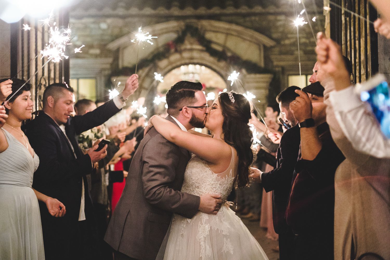 Bride and groom share romantic kiss surrounded by guests holding sparklers at wedding reception.