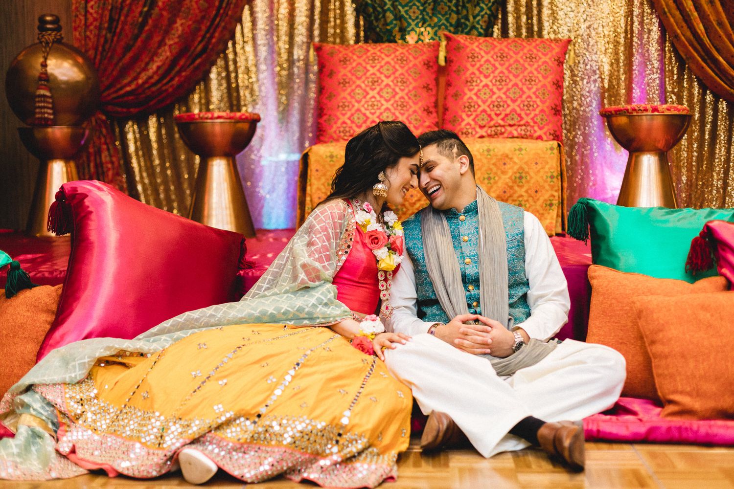 South Asian couple in yellow and teal wedding attire laughing together on colorful cushioned seating.