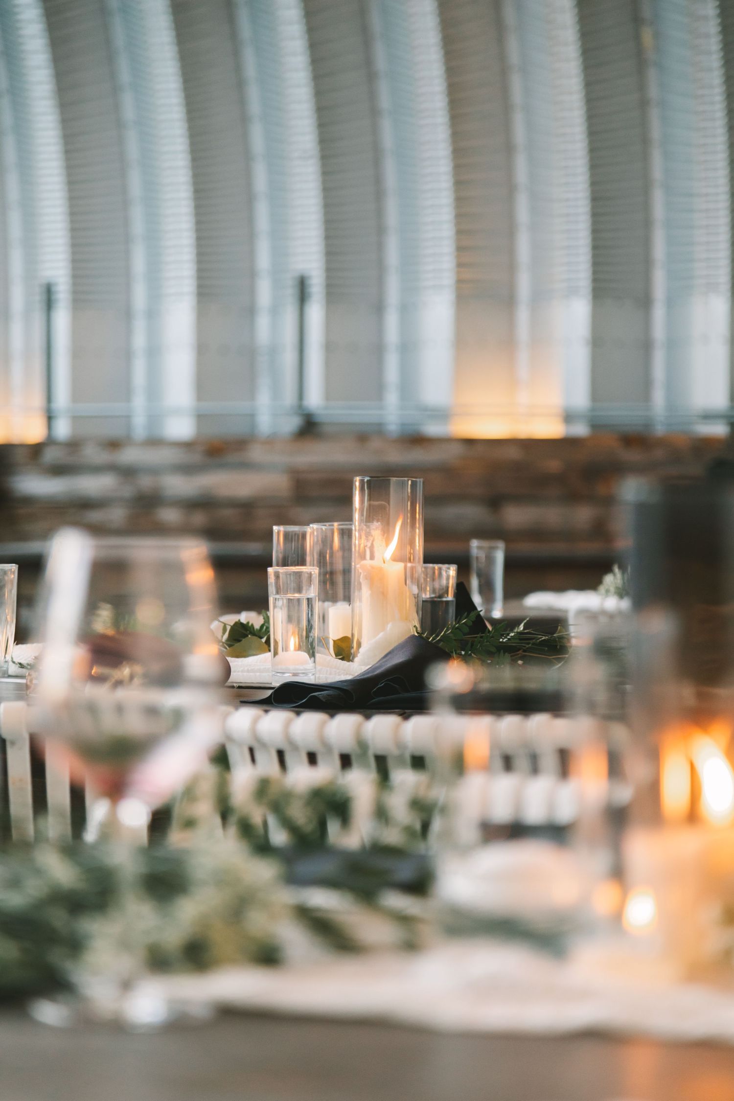 Elegant wedding reception table with glowing pillar candles and greenery garland in warmly lit venue.
