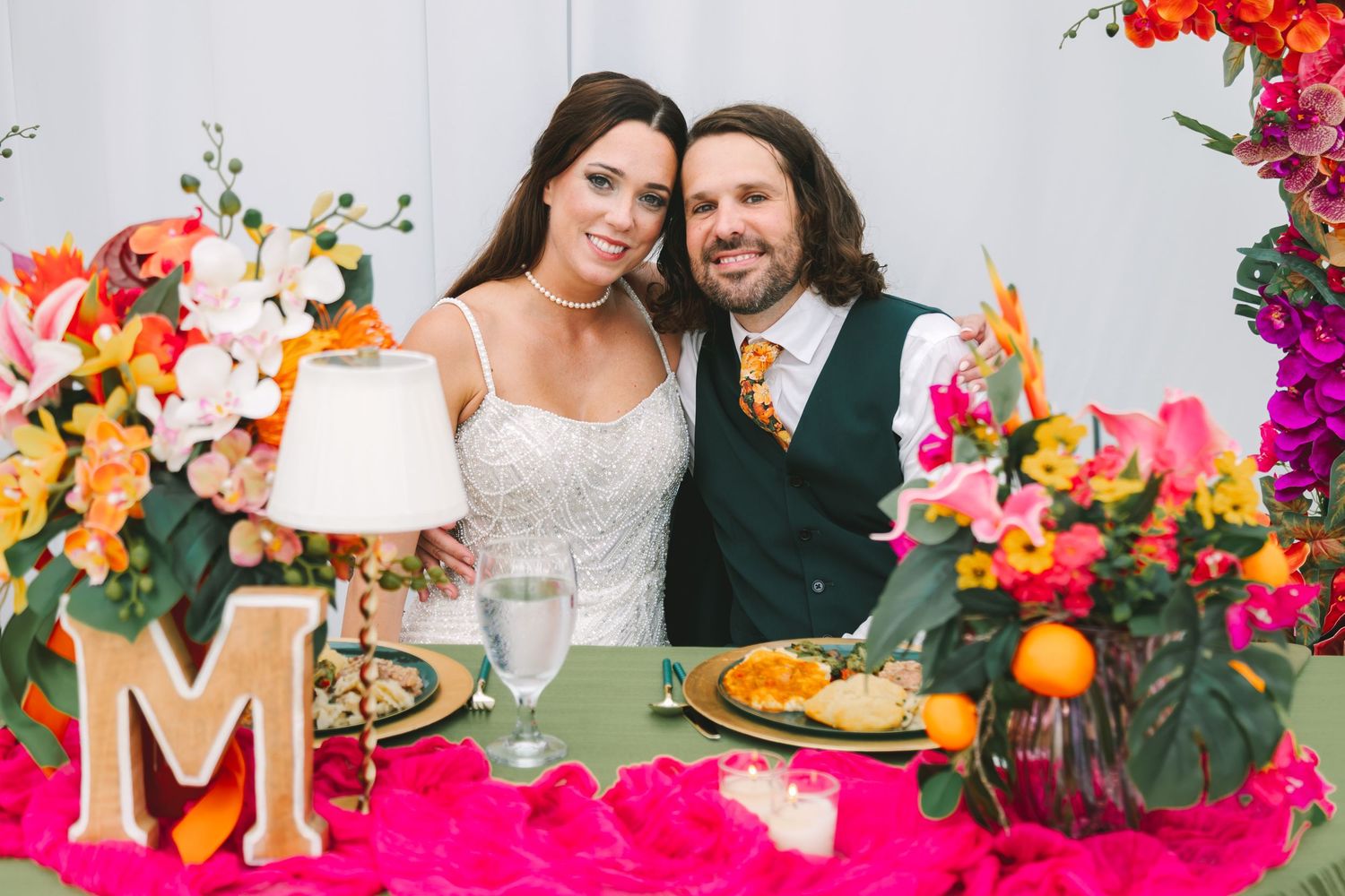 Smiling bride and groom seated at sweetheart table decorated with vibrant tropical floral arrangements.