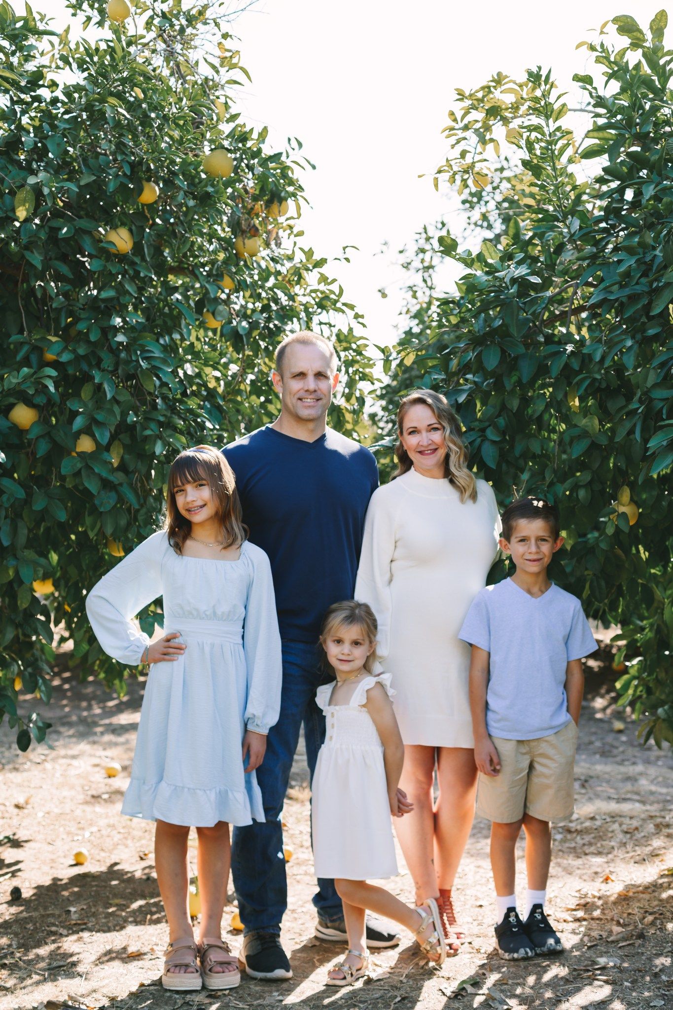 Happy family of five posing together in a sunny citrus orchard with lemon trees in background.