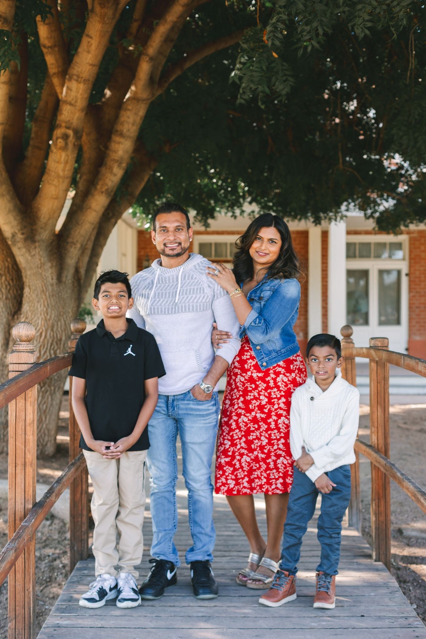 Hispanic family of four stands together on wooden bridge under large shade tree.
