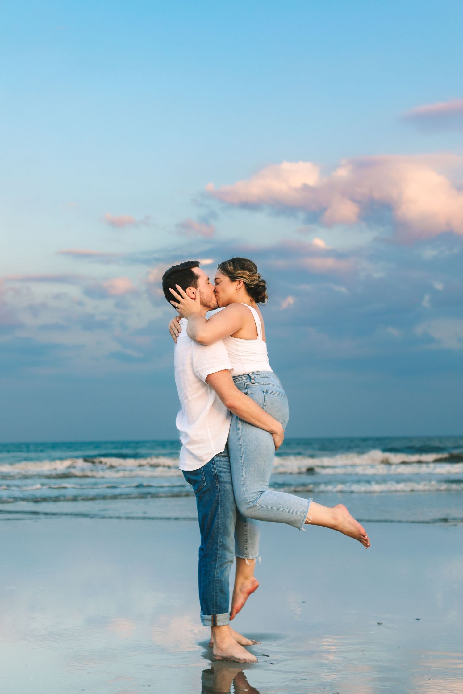 Couple kissing on the beach at sunset, man lifting woman in white top and jeans.