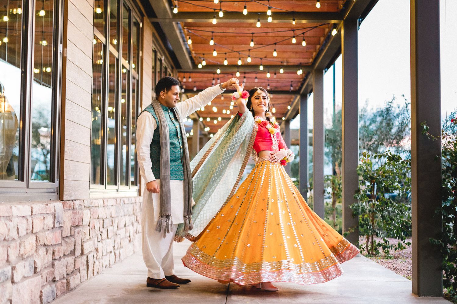 Indian groom twirling bride in vibrant yellow lehenga under string lights at outdoor wedding venue.