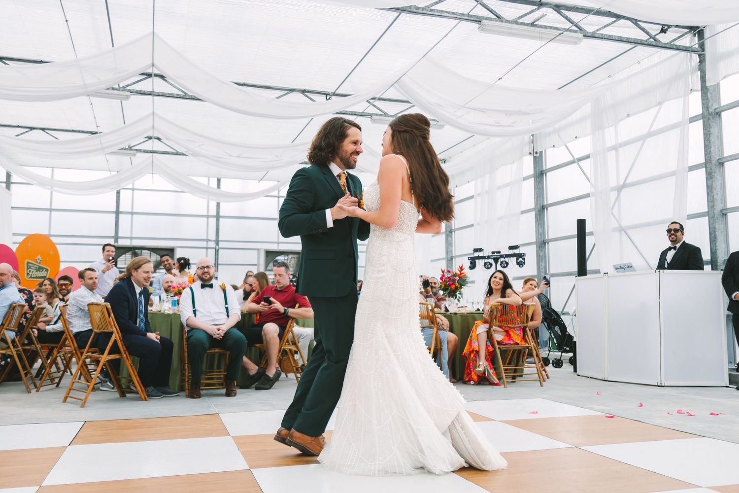 Bride and groom share first dance on checkered floor inside tent with seated wedding guests.