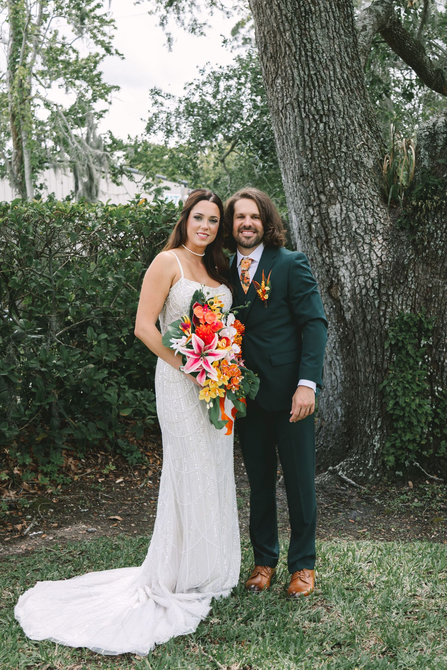 Bride and groom posing outdoors near a large tree, bride holding vibrant tropical bouquet, groom in green suit.