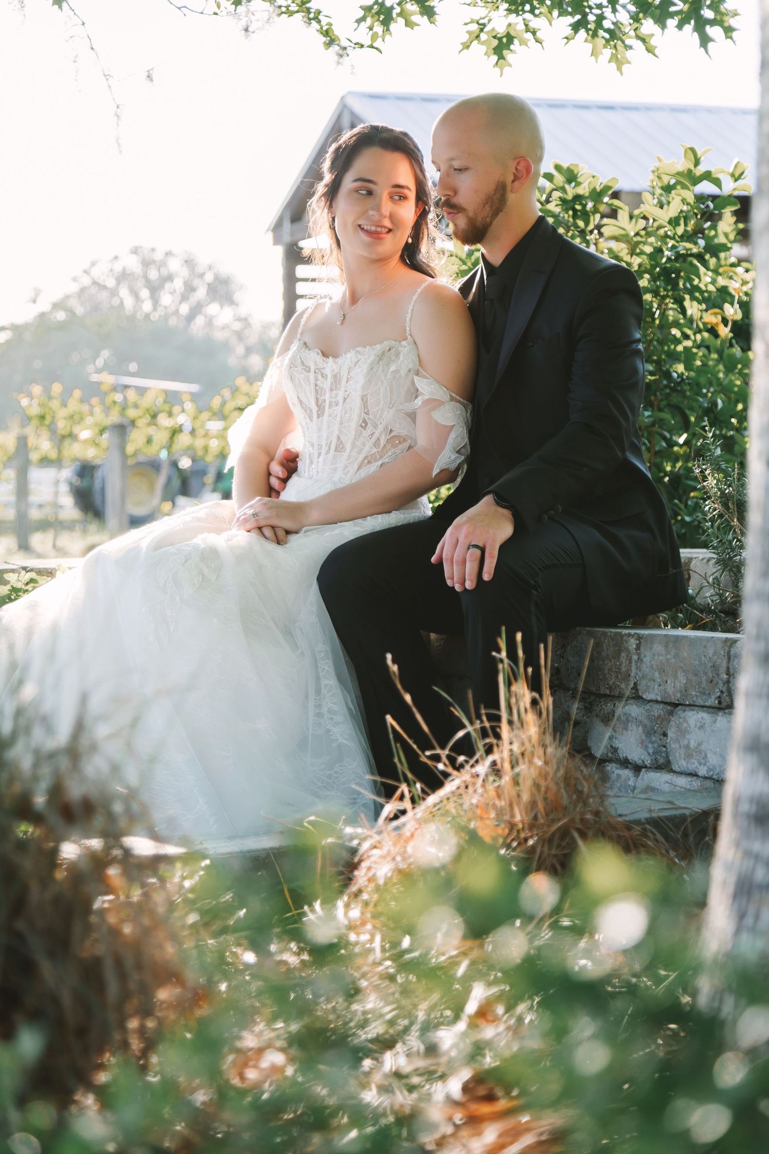 Bride and groom sitting together on stone steps surrounded by lush greenery in golden hour sunlight.