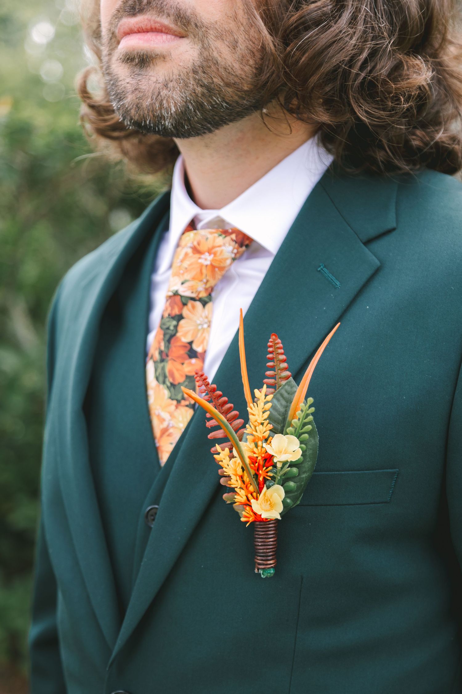 Groom in green suit with floral tie and feather boutonniere at wedding ceremony.