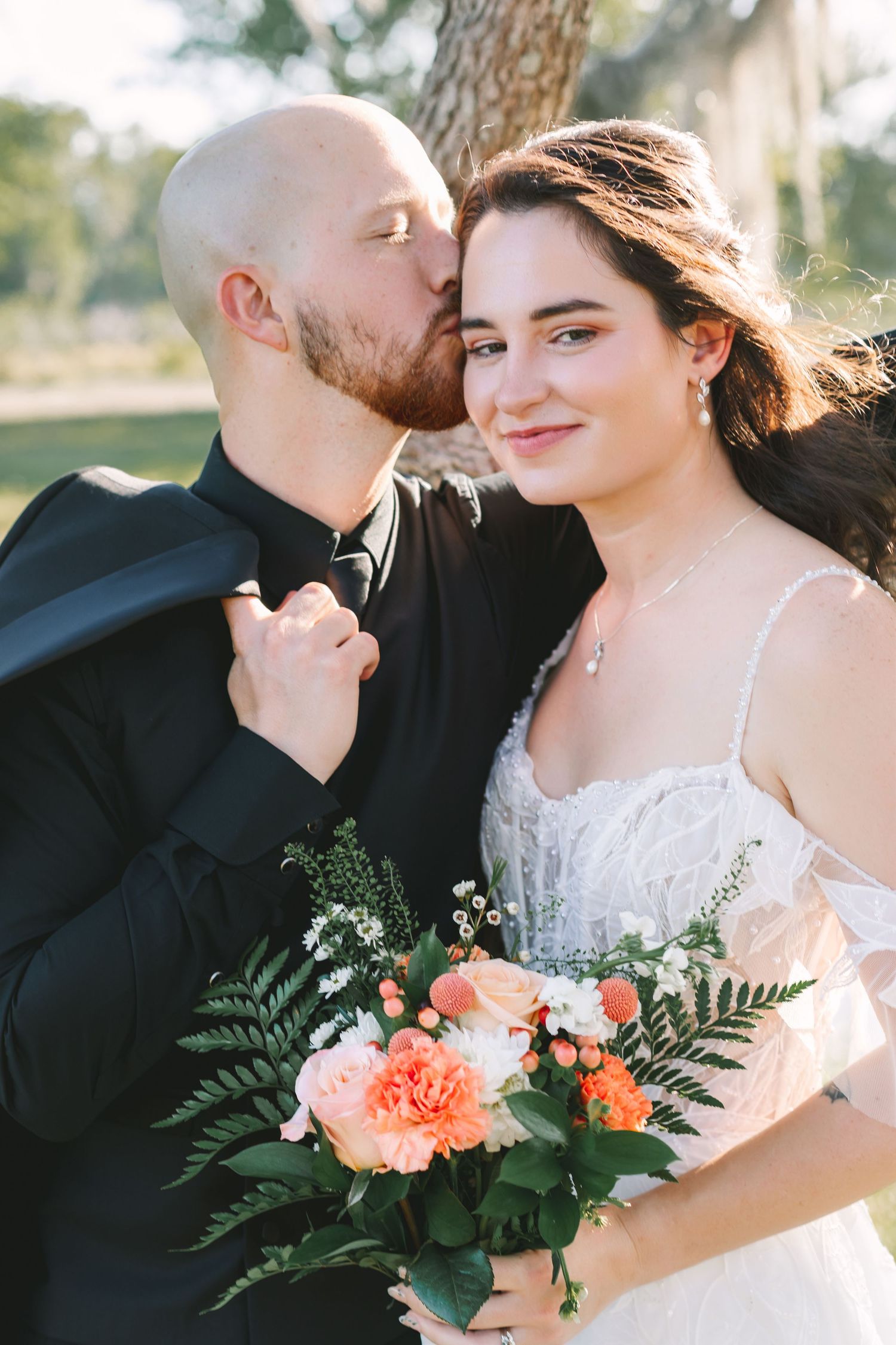 Bald groom in black suit kisses bride's cheek as she smiles holding peach floral bouquet.
