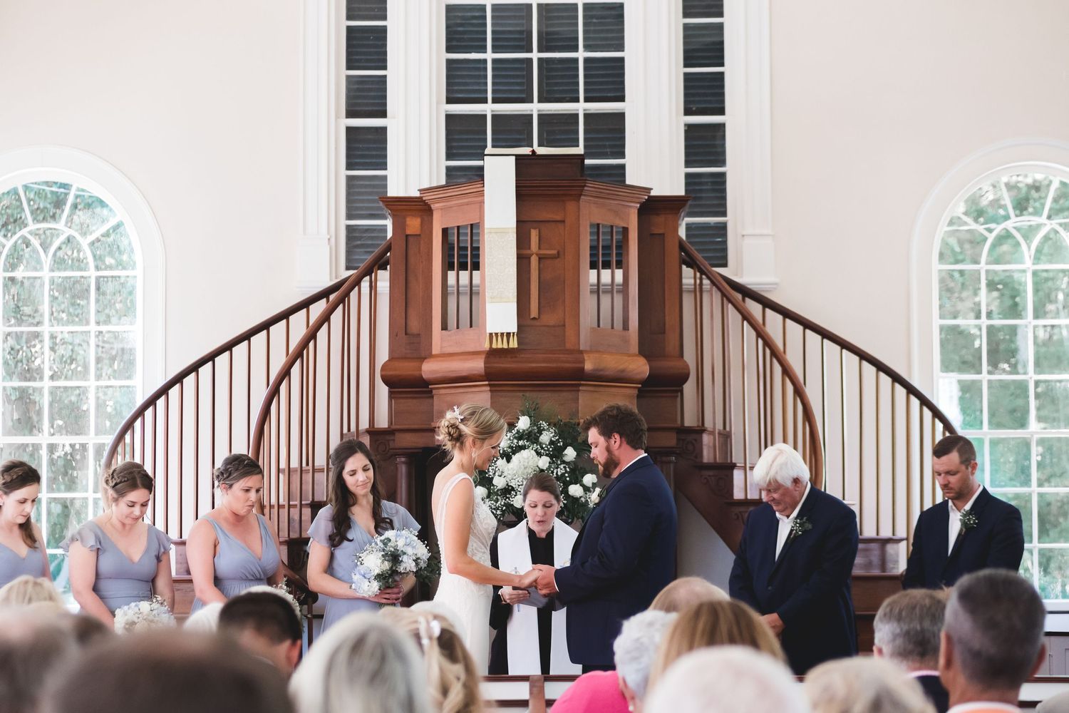 Bride and groom exchange vows at classic white church altar with curved staircases.