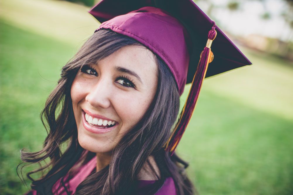 Happy young woman in maroon graduation cap and gown smiling on green grass.
