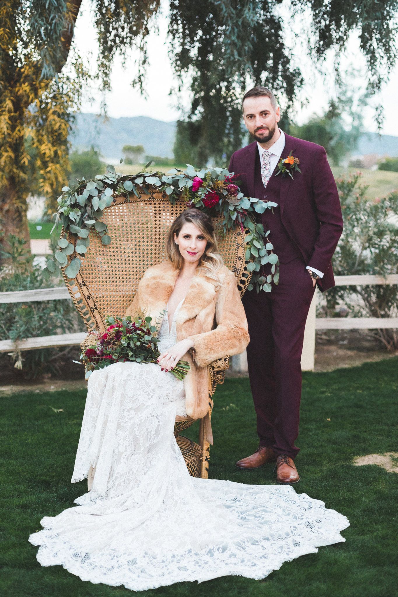 Bohemian bride and groom posing with floral peacock chair in outdoor wedding setting.