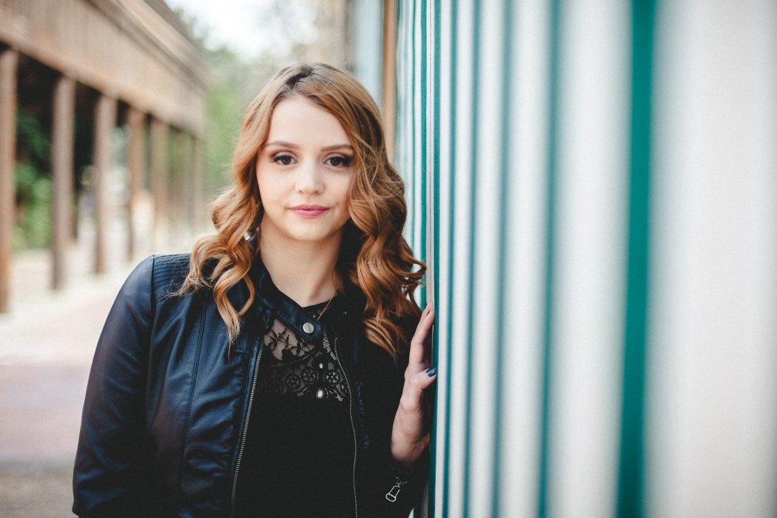 Teen girl in leather jacket leaning against colorful striped wall in urban setting.