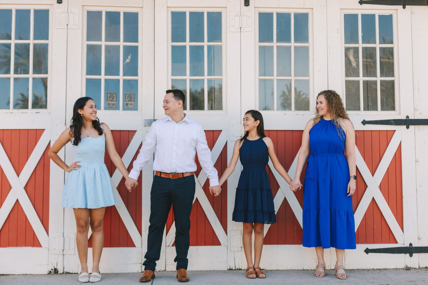 Four adults holding hands in front of a white barn with red doors on a sunny day.