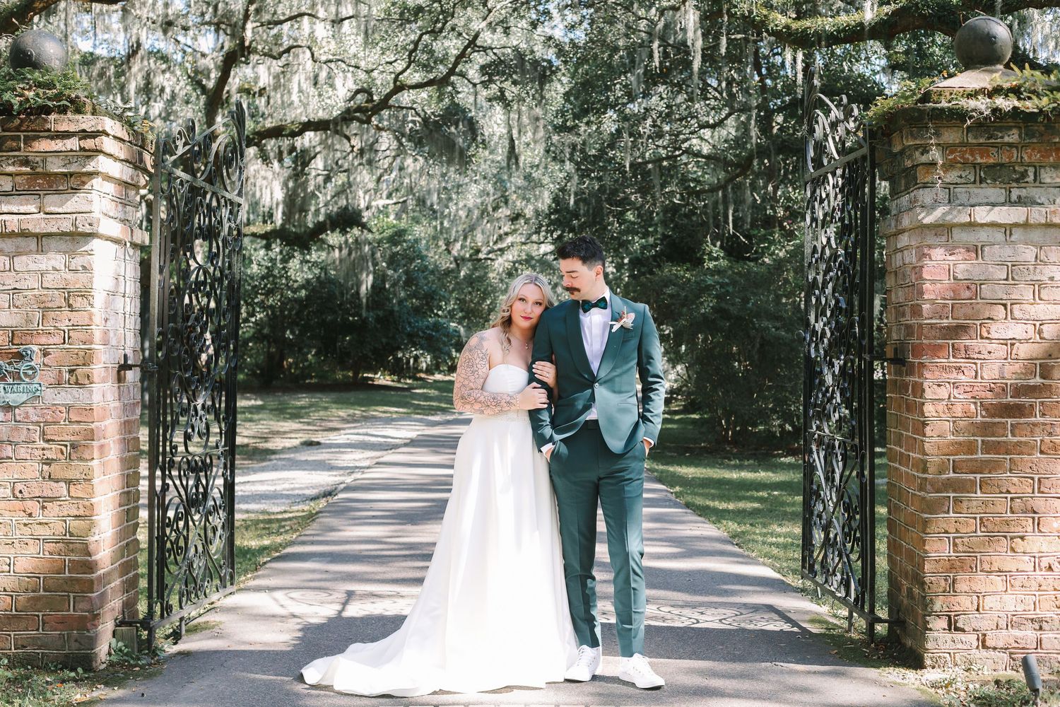 Bride and groom share a romantic moment under moss-draped trees at elegant iron garden gates.