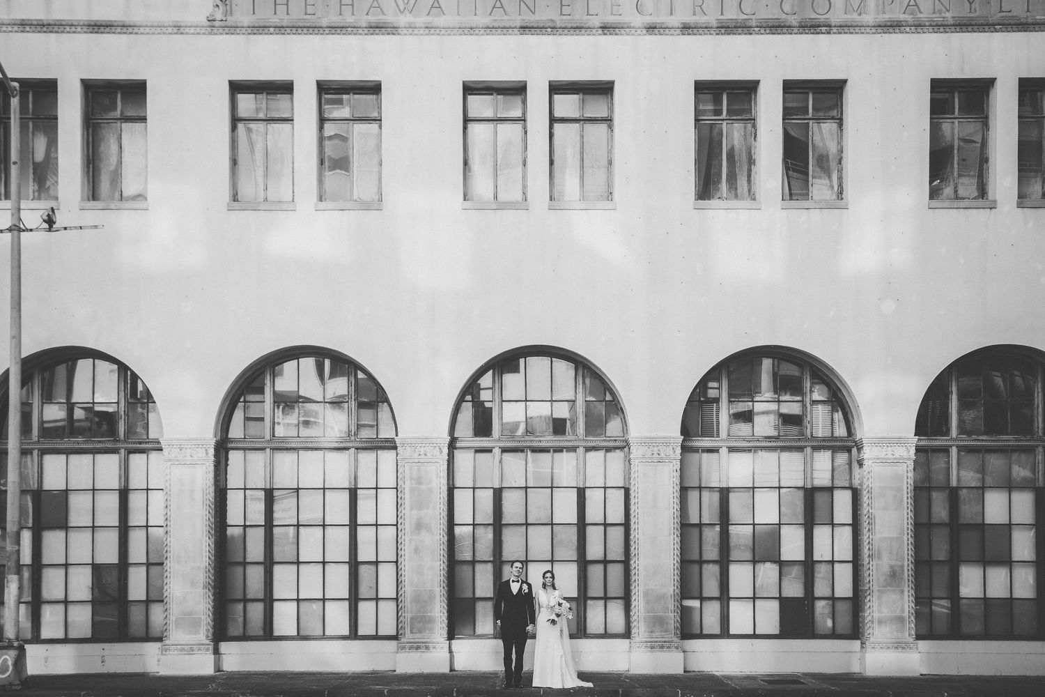 Black and white wedding photo of bride and groom standing before grand arched windows of The Hawaiian building.