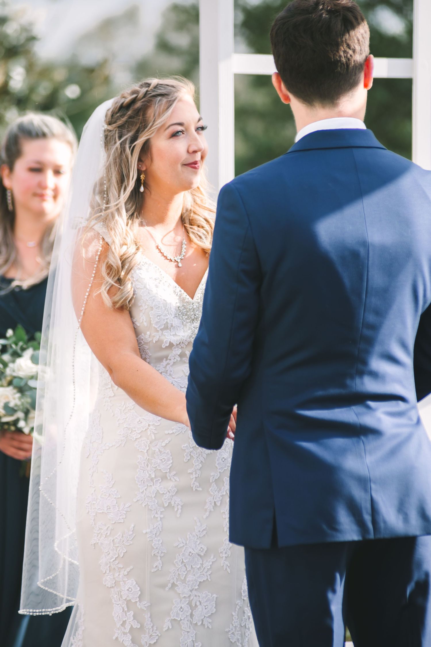Bride in lace gown and veil smiles emotionally at groom during outdoor wedding ceremony.