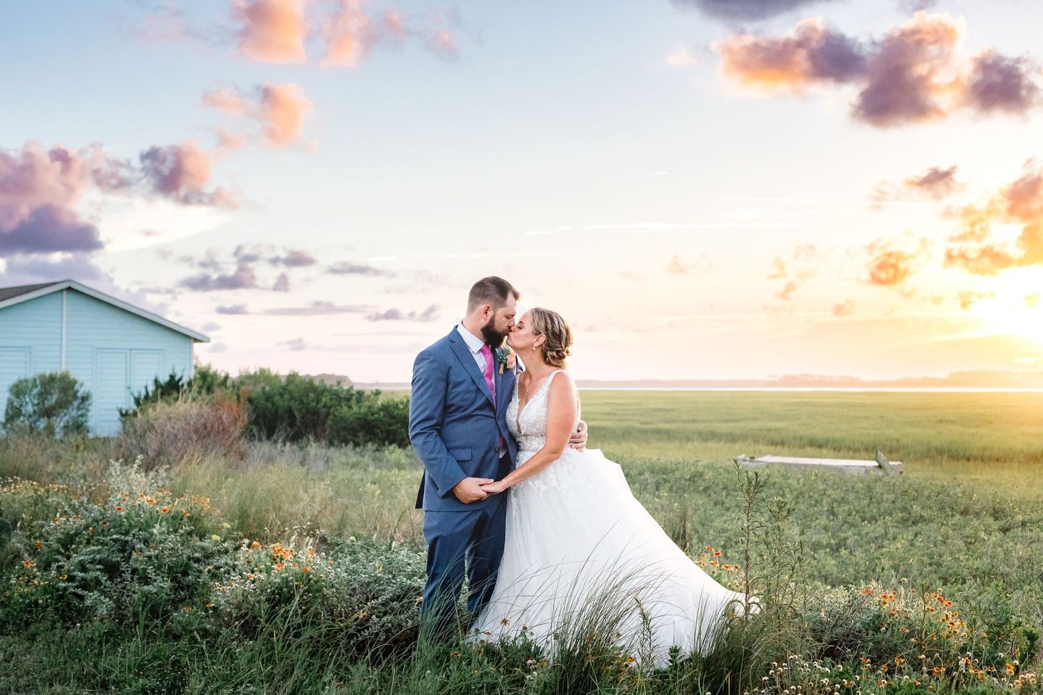 Bride and groom sharing a kiss in a green meadow beneath a stunning colorful sunset sky at golden hour.