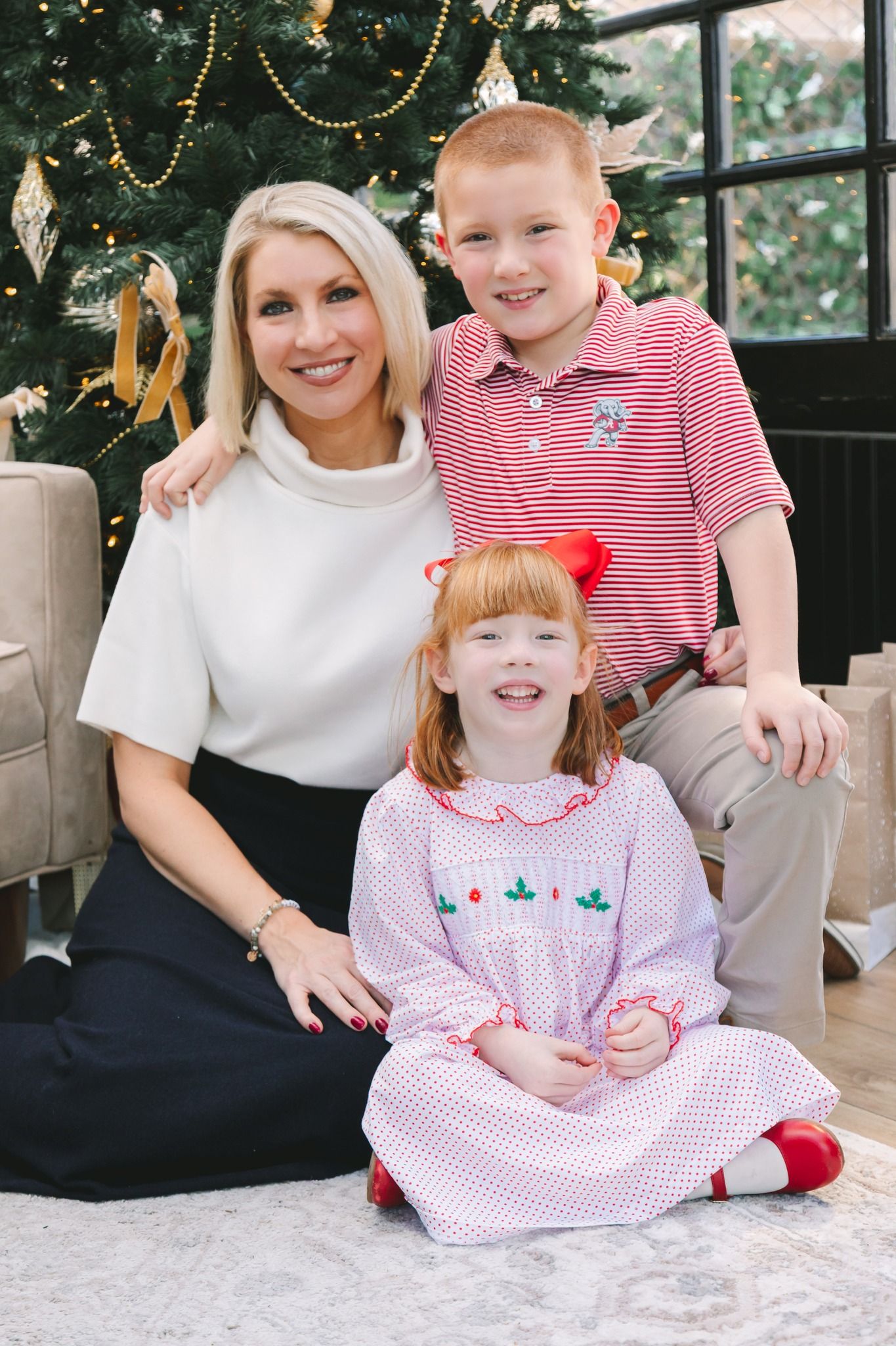 Smiling mother posing with two red-haired children in front of a decorated Christmas tree.