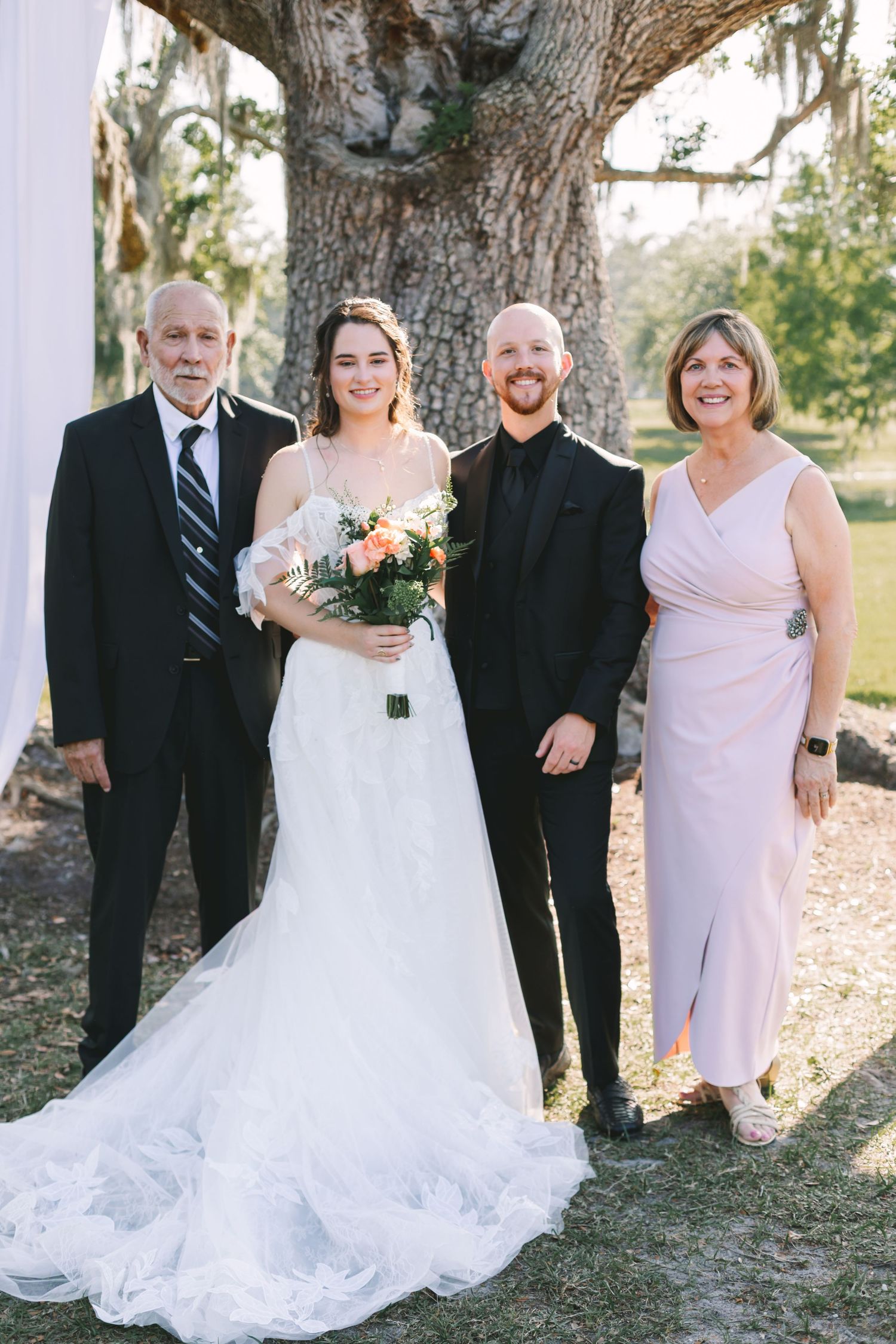 Bride and groom pose with parents under a large oak tree at an outdoor Florida wedding ceremony.